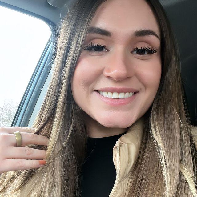 Woman smiling wearing a gold smart ring, beige jacket, and black shirt.