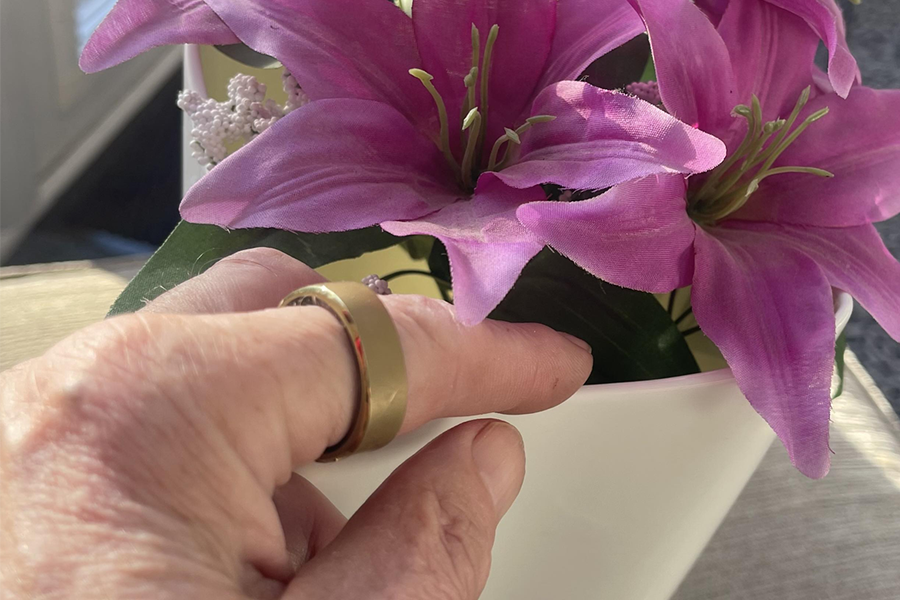 Gold smart ring worn on a finger next to pink artificial lilies, showcasing the ring's design and finish